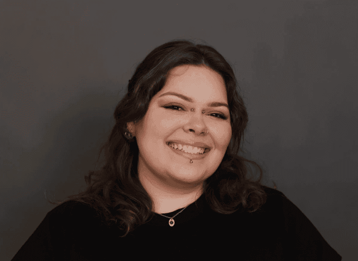 Woman smiling confidently against a dark background, with makeup and piercings visible.